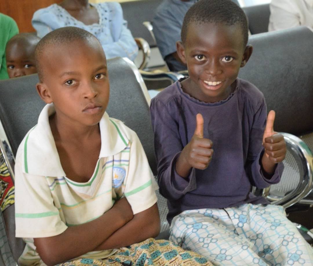 Two children seated in a waiting room, one smiling with both thumbs up